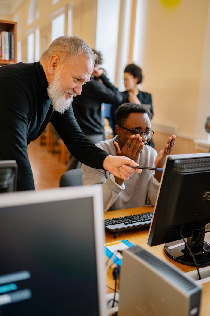 Elderly man guiding young adult on computer in office setting, showcasing mentorship and learning.